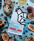 Rebel Dog coffee package surrounded by blueberries, passion fruits, and a teacup on a light blue background.
