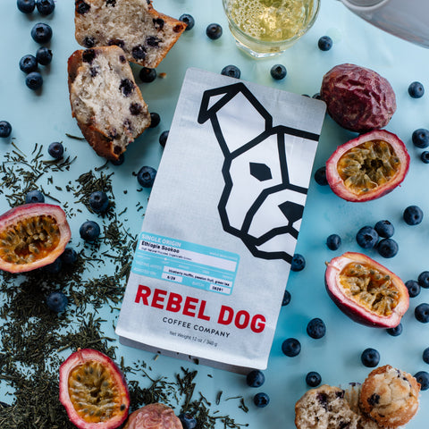 Rebel Dog coffee package surrounded by blueberries, passion fruits, and a teacup on a light blue background.
